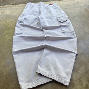 Pode incluir: Calças cargo bege claro com dois bolsos traseiros grandes, dois bolsos laterais tipo cargo com abas e uma pequena etiqueta retangular da marca no cós traseiro. As calças têm pernas largas, costuras visíveis ao longo das pernas e uma etiqueta lateral.