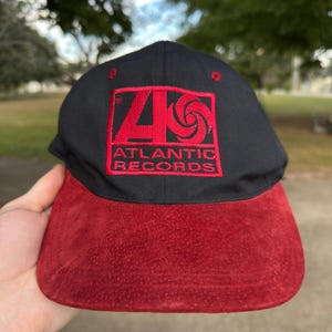 May include: A black baseball cap with a red suede brim and a red embroidered Atlantic Records logo on the front. The logo features the stylized letter A and a circular graphic above the text Atlantic Records, all centered on the black crown.