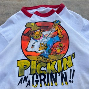Könnte beinhalten: Ein weißes Ringer-T-Shirt mit rotem Saum an Kragen und Ärmeln. Die Grafik zeigt eine Cartoon-Figur, die in einem rot-orangen Kreis Banjo spielt, daneben ein kleines Huhn und der aufgedruckte Text "PICKIN' AN A GRIN'N!!" unter dem Motiv.