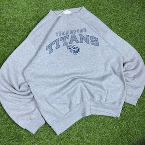 Op de afbeelding: Een grijs gemêleerd sweatshirt met lange mouwen en ronde hals, voorzien van de tekst Tennessee Titans in donkerblauwe letters gebogen boven het teamlogo, bestaande uit een gestileerd blauw en wit vlamontwerp rond een letter T.