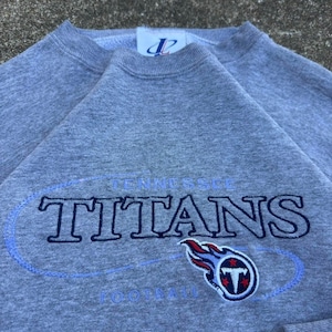 Op de afbeelding: Een grijs gemêleerd sweatshirt met ronde hals en het geborduurde opschrift Tennessee Titans in donkerblauw met een lichtblauwe omlijning. Onder de tekst staat het logo van de Tennessee Titans met een vlammend zwaard en een schild met een T.