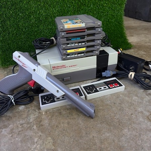 May include: A grey and white Nintendo Entertainment System console with two rectangular controllers, a grey Zapper light gun, a black power adapter, and a stack of five grey video game cartridges including Super Mario Bros and Duck Hunt.