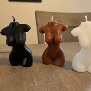 May include: Three torso-shaped candles in black, brown, and white. The candles are all the same size and shape, and they are arranged in a row on a wooden surface.
