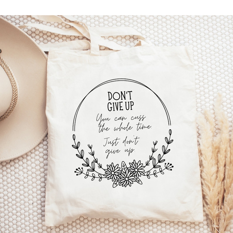 Puede incluir: Bolso tote de color crema con el texto "DON'T GIVE UP" en un dise&ntilde;o circular negro con detalles florales. La bolsa tambi&eacute;n incluye el texto "You can cuss the whole time. Just don't give up."