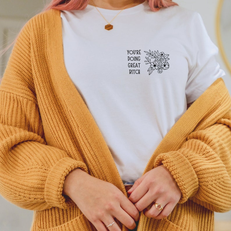 Puede incluir: Camiseta blanca con el texto "YOU'RE DOING GREAT BITCH" y un dise&ntilde;o floral negro. La camiseta se usa con una rebeca amarillo mostaza. Tambi&eacute;n se ven un collar y anillos dorados.