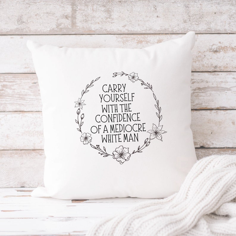 May include: White square cushion featuring the text "CARRY YOURSELF WITH THE CONFIDENCE OF A MEDIOCRE WHITE MAN" within a floral wreath. The cushion rests on a light wood surface with a white knitted blanket.