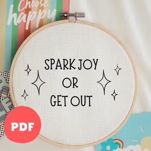 以下が含まれることがあります： 白い刺繍枠に黒い文字で「Spark Joy or Get Out」と書かれています。文字の周りに小さな星が4つあります。枠は白い表面の上に置かれています。