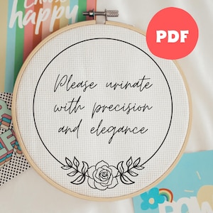 Peut inclure: Un cercle de broderie au point de croix avec le texte "Please urinate with precision and elegance" en écriture noire. Le motif comprend un élément floral en bas. Un cercle rouge avec "PDF" est dans le coin supérieur droit.
