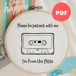 Puede incluir: Un bastidor de bordado de punto de cruz con el texto "Please be patient with me, I'm from the 1900s" y un diseño de casete. El bastidor se encuentra sobre un fondo de tarjetas coloridas y un círculo rojo "PDF".