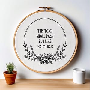 May include: A white embroidery hoop with black embroidery that reads "This too shall pass but like holy fuck" with a floral design around the text.