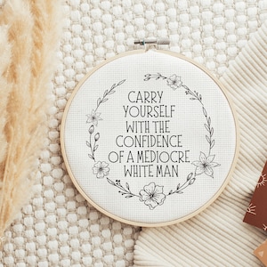 Puede incluir: Un bastidor de punto de cruz redondo y blanquecino con el texto en negro "CARRY YOURSELF WITH THE CONFIDENCE OF A MEDIOCRE WHITE MAN". El texto est&aacute; rodeado por una corona floral. Fondo neutro.