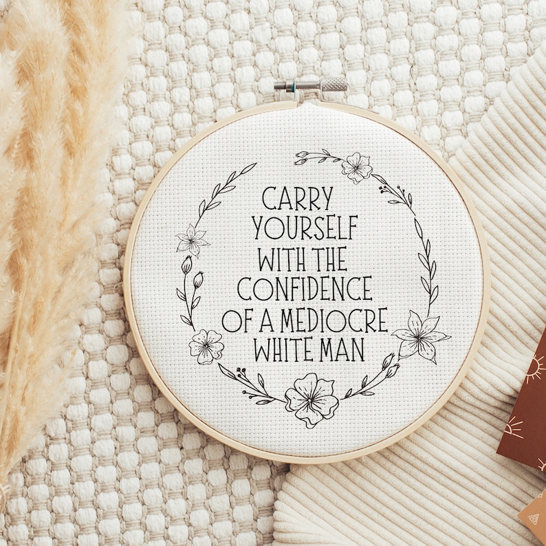 Puede incluir: Un bastidor de punto de cruz redondo y blanquecino con el texto en negro "CARRY YOURSELF WITH THE CONFIDENCE OF A MEDIOCRE WHITE MAN". El texto est&aacute; rodeado por una corona floral. Fondo neutro.