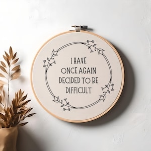 May include: A white embroidery hoop with a black floral wreath and the text "I have once again decided to be difficult".