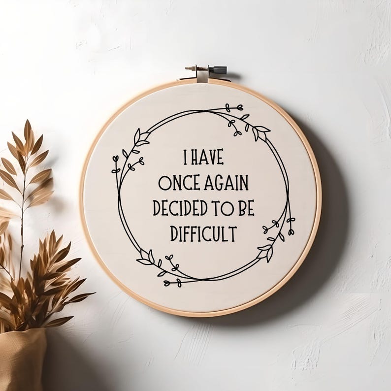 May include: A white embroidery hoop with a black floral wreath and the text "I have once again decided to be difficult".