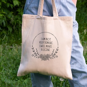 May include: A white canvas tote bag with black text that reads "I am not very demure I will make a scene" surrounded by a floral wreath.