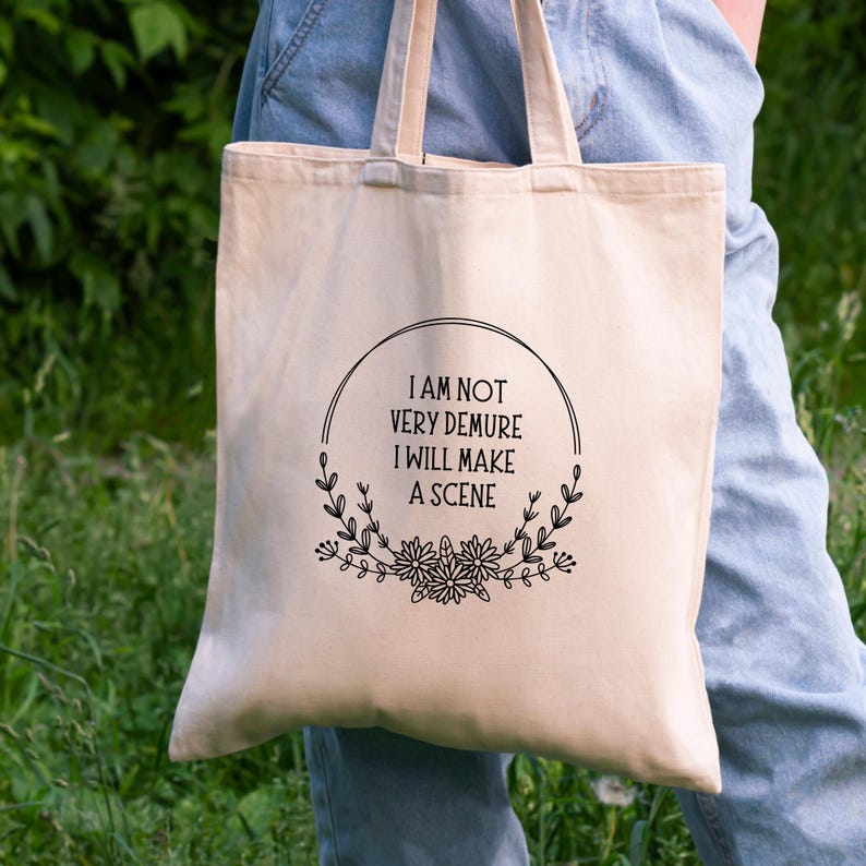 May include: A white canvas tote bag with black text that reads "I am not very demure I will make a scene" surrounded by a floral wreath.