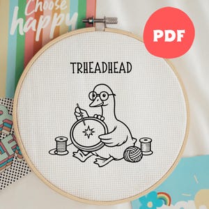 May include: A cross-stitch hoop with a cartoon duck wearing glasses, stitching. The text "THREADHEAD" is above the duck. The hoop is surrounded by spools of thread and a ball of yarn. A red circle with "PDF" is in the upper right corner.