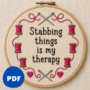 May include: A circular wooden embroidery hoop containing off-white fabric with the black embroidered text Stabbing things is my therapy. The design features six brown spools with pink thread, two pairs of crossed silver needles, and two pink hearts.