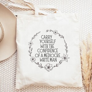 May include: A cream-coloured tote bag featuring the text "CARRY YOURSELF WITH THE CONFIDENCE OF A MEDIOCRE WHITE MAN" within a black floral wreath. The bag has handles and is made of a light-coloured fabric.