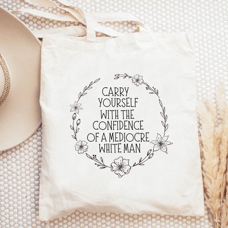 May include: A cream-coloured tote bag featuring the text "CARRY YOURSELF WITH THE CONFIDENCE OF A MEDIOCRE WHITE MAN" within a black floral wreath. The bag has handles and is made of a light-coloured fabric.