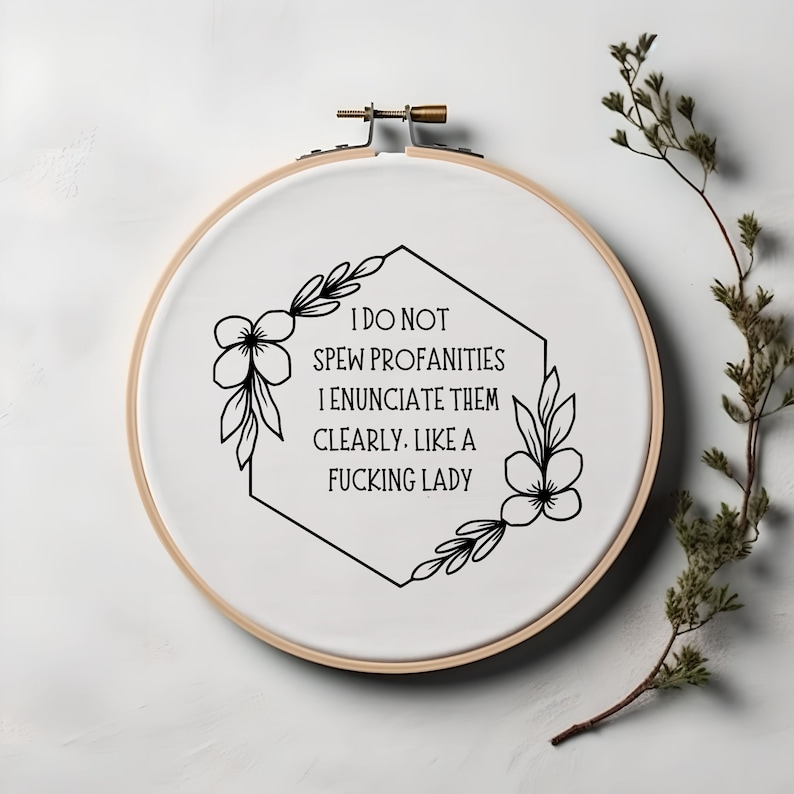 Puede incluir: Un bastidor de bordado de tela blanca con el texto "I DO NOT SPEW PROFANITIES I ENUNCIATE THEM CLEARLY. LIKE A FUCKING LADY" en negro. El dise&ntilde;o incluye detalles florales y de hojas. Una ramita de vegetaci&oacute;n est&aacute; en el fondo.