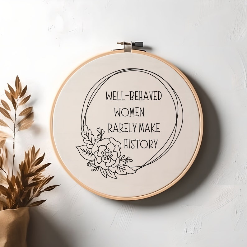 Puede incluir: Un bastidor de bordado redondo con el texto negro "WELL-BEHAVED WOMEN RARELY MAKE HISTORY". El dise&ntilde;o incluye una ilustraci&oacute;n floral y de hojas. El bastidor es de madera y la tela es de color beige claro.