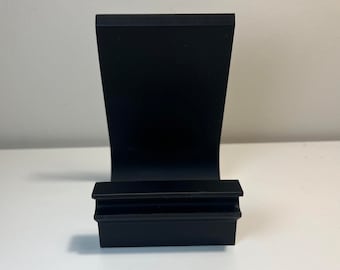May include: A black metal phone stand with a curved back and a rectangular base. The stand is designed to hold a phone or tablet upright. The stand is made of a solid, matte black metal.