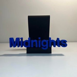 以下が含まれることがあります： 黒いスタンドに、青色の3Dプリント文字で「Midnights」と書かれています。文字はスタンドの前に配置され、レイヤー効果を生み出しています。背景はプレーンな白い表面です。