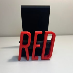 May include: A red 3D-printed word "RED" is displayed in front of a black rectangular stand. The word is in a bold, sans-serif font. The background is a plain white surface.