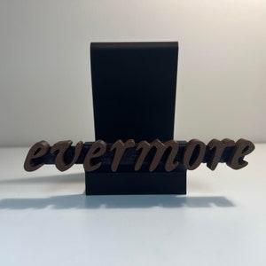 以下が含まれることがあります： 筆記体の「evermore」という文字が書かれた茶色の木製サイン。黒いベースに取り付けられ、黒い長方形の背景があります。白い表面に表示されています。