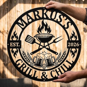 May include: A circular black metal wall sign featuring the text MARKUS'S at the top and GRILL & CHILL at the bottom. The center displays a grill with flames, a spatula, a fork, two beer mugs, and wheat stalks. The text EST. 2026 is on the sides.