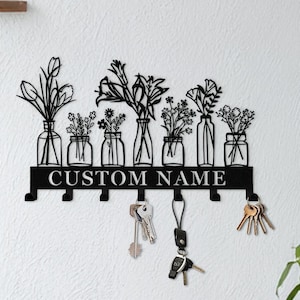 Könnte beinhalten: Schwarzer Metall-Schlüsselhalter mit floralem Design mit sechs Vasen mit verschiedenen Blumenarrangements. Der Schlüsselhalter hat Haken darunter und den Text "CUSTOM NAME" in der Mitte. Schlüssel hängen an einigen Haken.