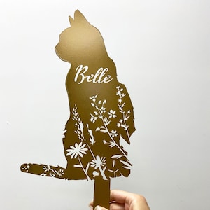 May include: A gold-colored metal cat silhouette garden stake with the name "Belle" in white script. The cat's body is filled with a white floral design. The stake is designed for outdoor use.