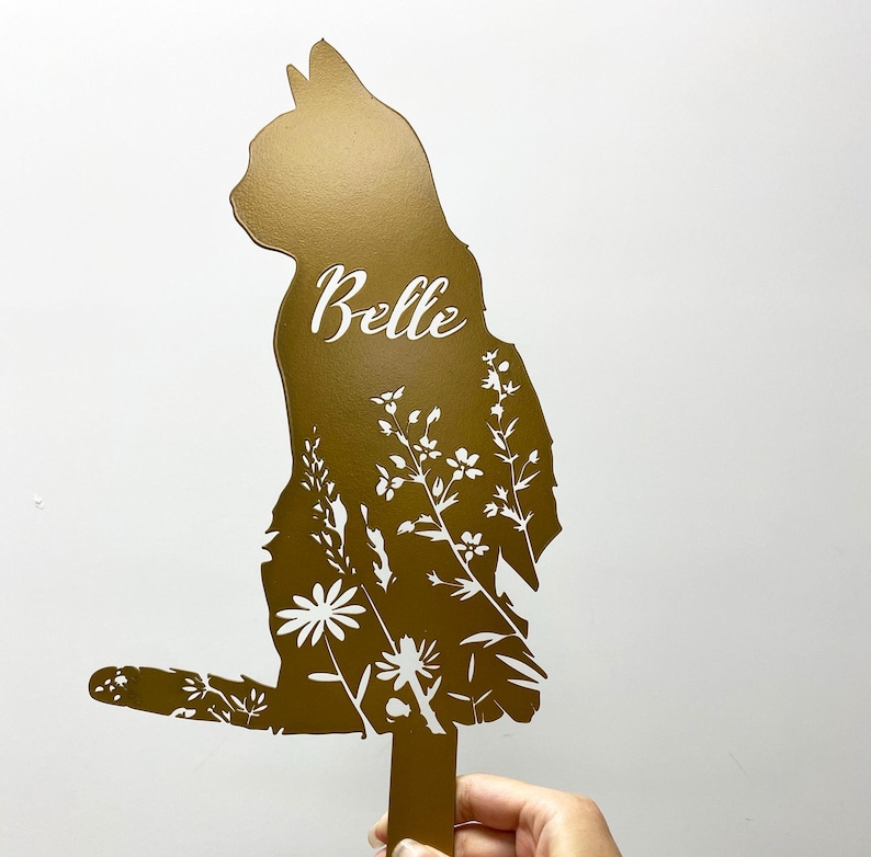 May include: A gold-colored metal cat silhouette garden stake with the name "Belle" in white script. The cat's body is filled with a white floral design. The stake is designed for outdoor use.