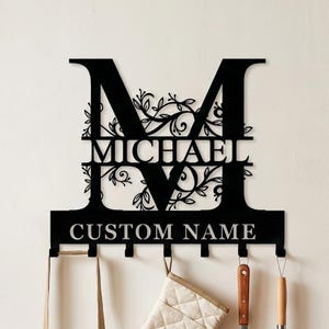 Könnte beinhalten: Schwarze Metall-Wanddekoration mit dem Buchstaben "M", dem Namen "MICHAEL" und "CUSTOM NAME" ausgeschnitten. Mit floralen Elementen und Haken für Küchenutensilien. Geeignet für Küche oder Essbereich.