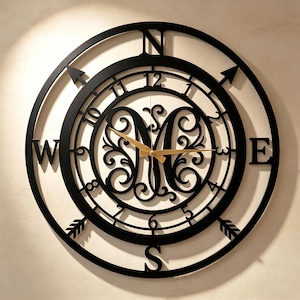 May include: A circular black metal wall clock featuring a compass design with cardinal directions N, S, E, and W. The center displays a large, ornate letter M surrounded by scrollwork. It has gold-colored clock hands and numbers 1 through 12.