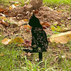 May include: Black metal cat silhouette garden stake with floral cutouts. The cat's name, "Milo", and the years "2010-2024" are inscribed on the cat's body. The stake is placed in a grassy area with fallen leaves.