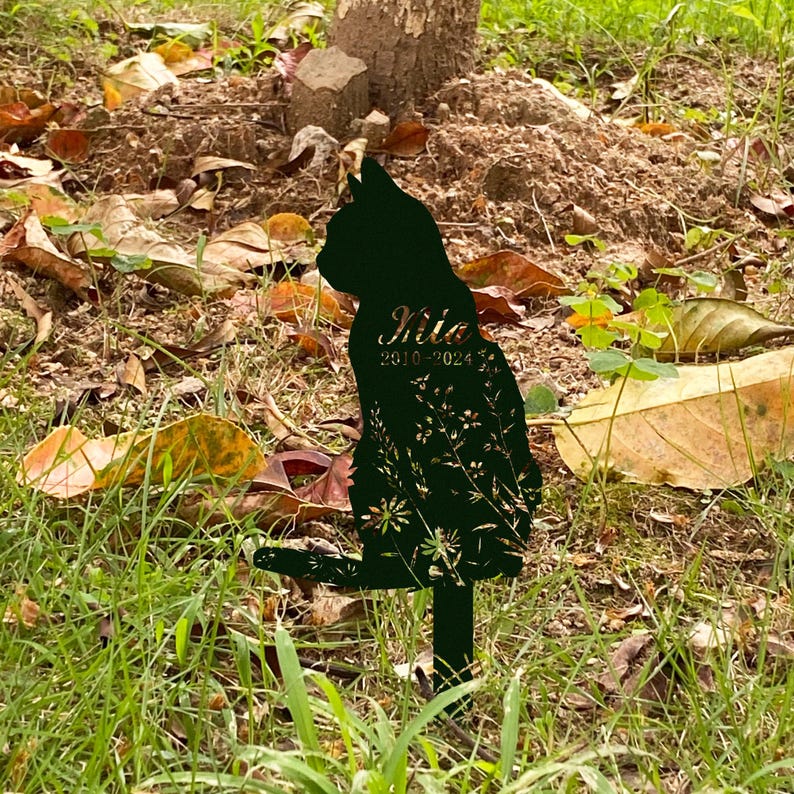 May include: Black metal cat silhouette garden stake with floral cutouts. The cat's name, "Milo", and the years "2010-2024" are inscribed on the cat's body. The stake is placed in a grassy area with fallen leaves.