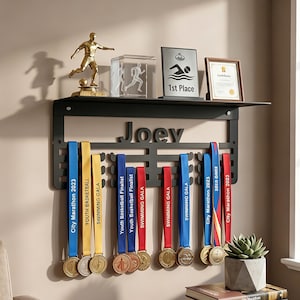 Puede incluir: Un expositor de medallas de pared en metal negro con el nombre Joey recortado. El estante superior sostiene un trofeo de fútbol dorado, un bloque de cristal, una placa de natación de primer lugar y un certificado, con medallas colgando debajo.