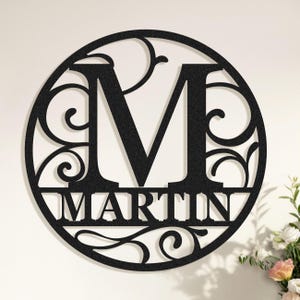 May include: Black metal wall art with the letter "M" and the name "MARTIN" in a circular design. The design includes decorative scrollwork. Ideal for home decor, this piece is a stylish addition.