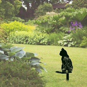 May include: Black metal cat garden stake with floral detailing. The cat silhouette has the name "Mia" and the years "2010-2024" inscribed on it. The stake is placed in a green grassy yard with lush foliage in the background.