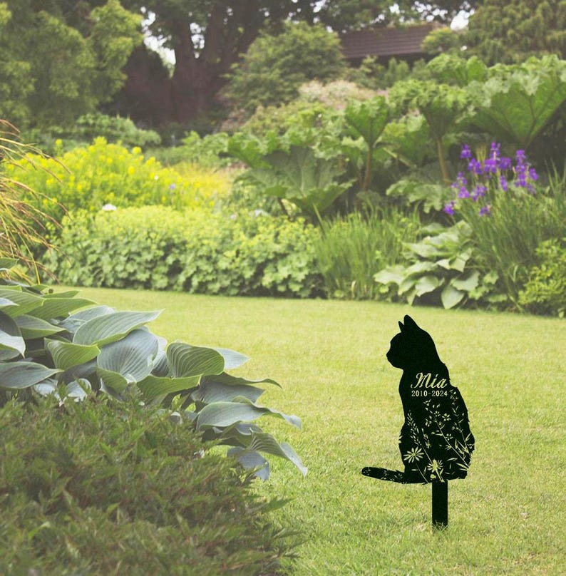 May include: Black metal cat garden stake with floral detailing. The cat silhouette has the name "Mia" and the years "2010-2024" inscribed on it. The stake is placed in a green grassy yard with lush foliage in the background.