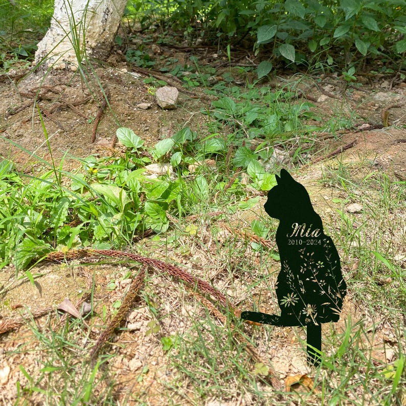 May include: A black metal cat silhouette garden stake. The cat is depicted in profile, with the name "Mia" and the years "2010-2024" etched on its side. The cat's body is filled with a floral design, and it is placed in a grassy outdoor setting.