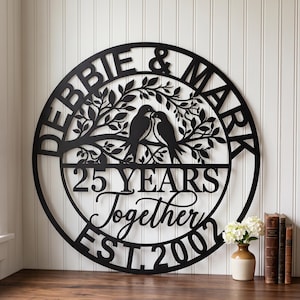 Op de afbeelding: Een rond zwart metalen wandbord met een uitgesneden ontwerp van twee vogels op een tak in een boom met bladeren. De tekst luidt DEBBIE & MARK, 25 YEARS Together en EST. 2007, gerangschikt in een cirkelvormig patroon rond het centrale vogelmotief.
