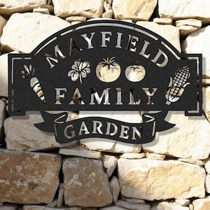 Op de afbeelding: Zwart metalen tuinbord met de woorden "MAYFIELD FAMILY GARDEN" uitgesneden. Het bord heeft uitgesneden afbeeldingen van een wortel, klaver, tomaten en maïs. Het bord is op een stenen muur gemonteerd.