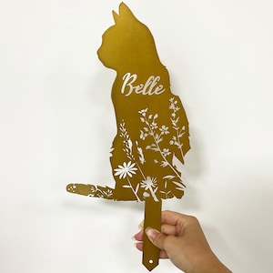 May include: A golden-colored cat-shaped garden stake with floral cutouts. The name "Belle" is written in cursive. The stake is held by a hand against a white background. The cat is in a sitting position.