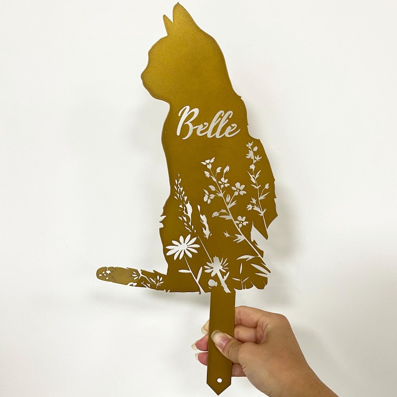 May include: A golden-colored cat-shaped garden stake with floral cutouts. The name "Belle" is written in cursive. The stake is held by a hand against a white background. The cat is in a sitting position.