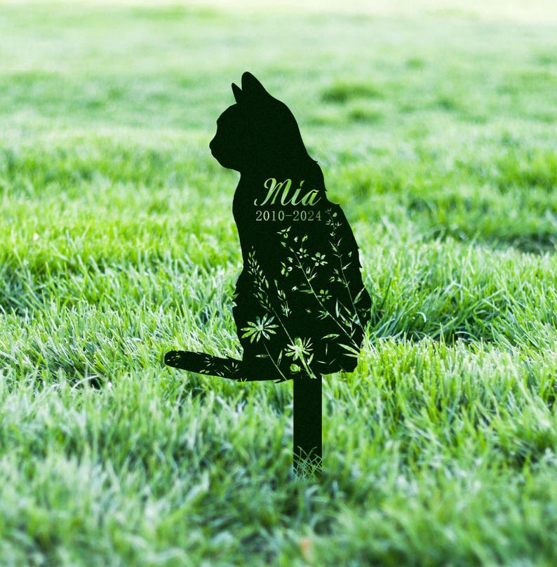 May include: Black metal cat silhouette garden stake with the name "Mia" and dates "2010-2024" etched on the front. The cat's body is filled with a floral design. The stake is placed in a grassy area.
