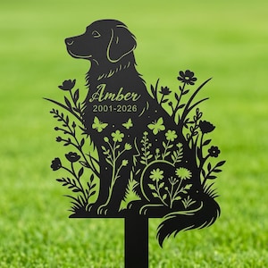 Op de afbeelding: Zwarte metalen tuinpaal met een silhouet van een hond, omringd door bloemen, vlinders en bladeren. De naam van de hond, "Amber", en de jaren "2001-2026" zijn gegraveerd. De paal is bedoeld om in de tuin te plaatsen.