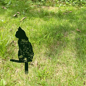 May include: A black metal cat silhouette garden stake with the name "Mia" and the years "2010-2024" etched on the front. The cat is decorated with floral designs and is placed in a grassy area.